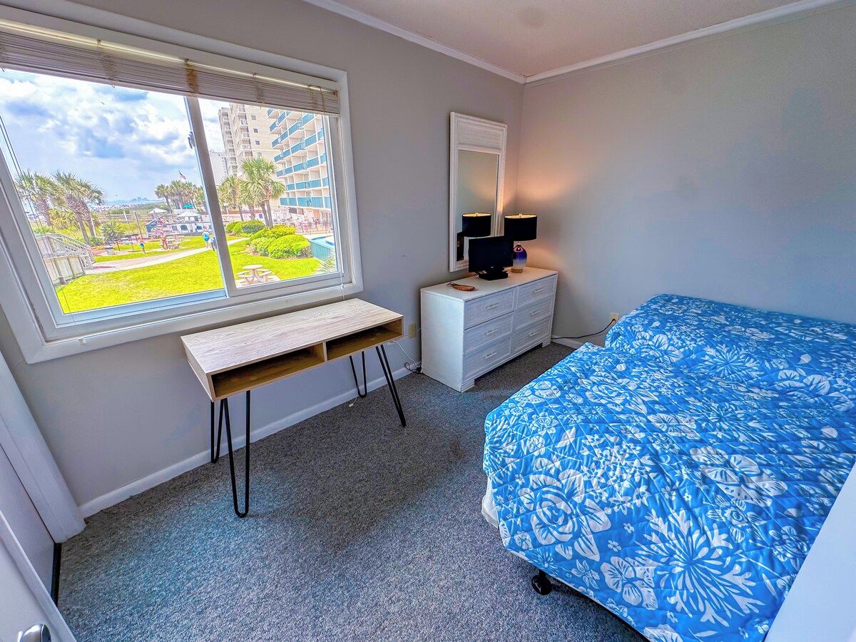 Ein Schlafzimmer mit Bett, Schreibtisch, Kommode, Spiegel und Fenster mit Blick auf ein Gebäude und Grünflächen.