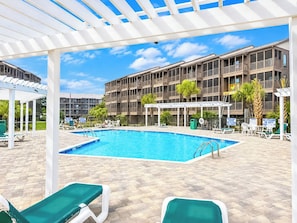 A swimming pool area with lounge chairs, a covered seating area, and a multi-story building in the background.