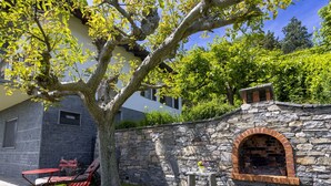 A stone wall with a brick oven, a tree, and a red table and chair set in a grassy area.