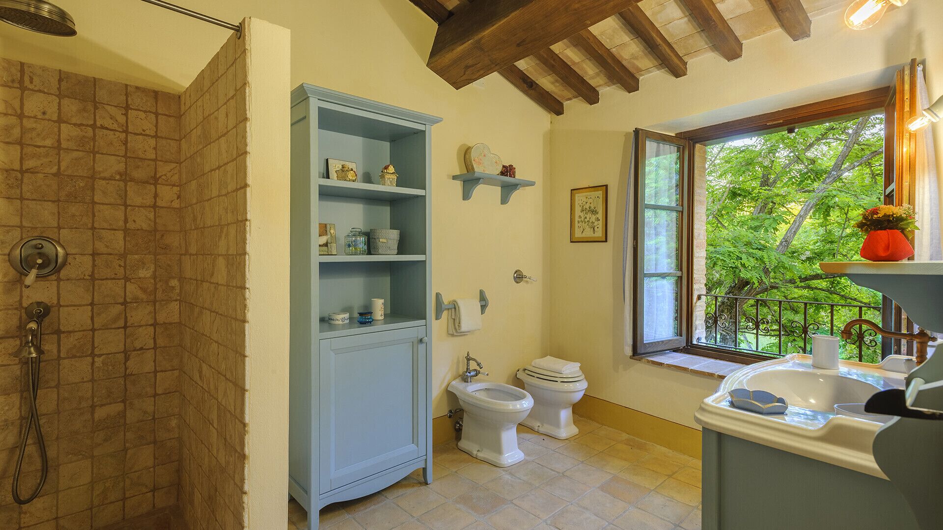 A bathroom with a shower, two toilets, a blue cabinet with shelves, a window with a view of greenery, and a sink with a vase of flowers.