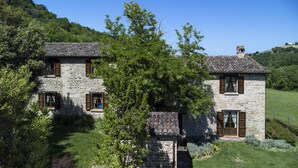 Una casa de piedra con techo de tejas, rodeada de exuberante vegetación y un paisaje montañoso.