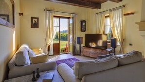 Una sala de estar con sofá, mesa de centro, televisor y chimenea. Hay una ventana con cortinas y vistas al exterior.