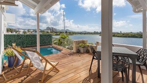 Eine Holzterrasse mit Pool, Liegestühlen und einem Tisch mit Stühlen, mit Blick auf ein Gewässer und entfernte Gebäude.