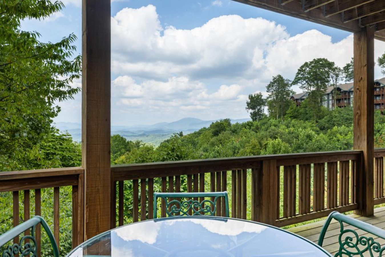 Une terrasse en bois avec une table et des chaises en verre, offrant une vue sur un paysage verdoyant et luxuriant ainsi que sur des montagnes lointaines.