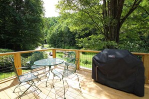 Eine Holzterrasse mit Tisch und Stühlen, umgeben von einem Hollerdgeschoss, mit Blick auf ein bewaldetes Gebiet.