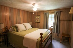 Ein Zimmer mit einer holzverkleideten Wand, einem Bett mit Holz-Kopfteil, einem Nachttisch mit Lampe und einem Fenster mit Vorhängen.
