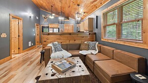 A cozy living room with a wooden ceiling, a large window with blinds, a sofa set, and a coffee table with books.