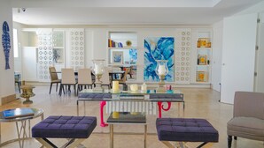 Una sala de estar moderna con una mesa de centro de vidrio, dos zonas de estar con cojines, un comedor con sillas y un cuadro abstracto grande en la pared.
