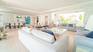 Amplia sala de estar con un sofá blanco grande, una mesa de centro de vidrio y un sillón gris. Hay una zona de comedor con una mesa y sillas blancas, y una cocina al fondo.