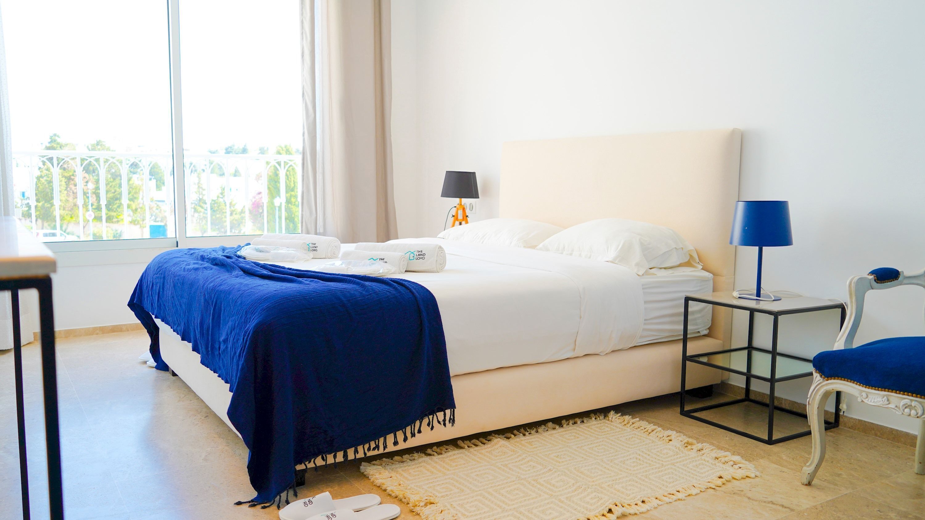 Una cama bien hecha con sábanas blancas y una manta azul, una mesita de noche con lámpara, una silla y vistas a un balcón con follaje.