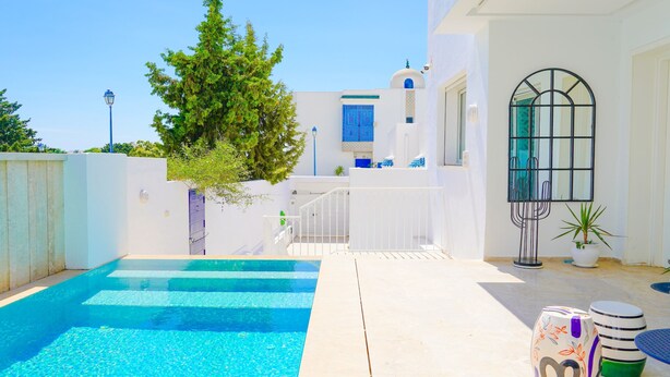 Un área de piscina con una piscina azul cristalina, paredes blancas y un árbol.