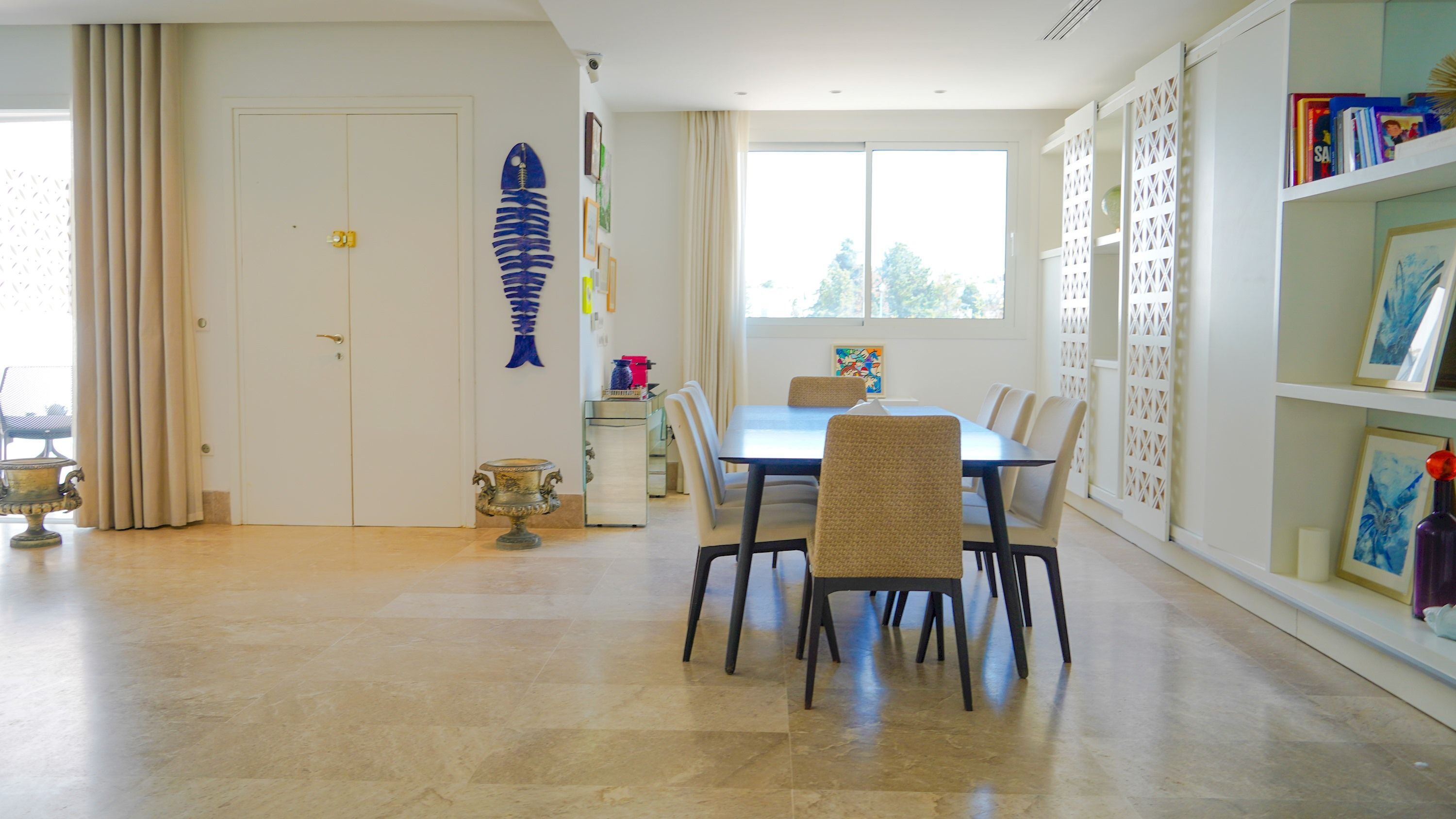Uma área de refeições com uma mesa e cadeiras, um esqueleto de peixe decorativo na parede, e uma estante com livros e um vaso.