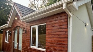 A wooden house with a gabled roof, a front door, and two windows with shutters. - 1-bed Cosy Cottage Near Emerald Park - Sleeps 5 (Rackenstown)