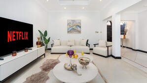 Ein modernes Wohnzimmer mit einem Flachbildfernseher, der 'NETFLIX' anzeigt, einem weißen Sofa, einem Couchtisch mit Dekoration und einer Deckenleuchte.