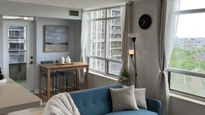 A modern living room with a blue sofa, a dining table with chairs, a large window with curtains, and a clock on the wall. - Premium 2BR Extended Stay Near YYZ Free Parking Included (Toronto)