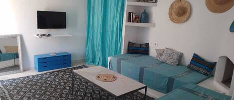 A room with a TV, two beds with blue bedding, a patterned rug, a blue dresser, and a window with curtains.