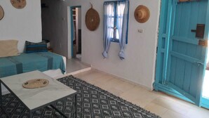 A room with a bed, a table, a patterned rug, a blue door, and a window with curtains. - Welcome to Studio Jasmin in this corner of paradise in Menzel Soltane Ahmed (MIDOUN)