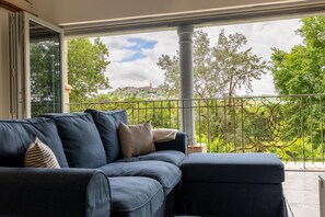 Un balcon avec un canapé et un pouf, une balustrade en fer forgé et une vue sur la verdure et une colline lointaine.