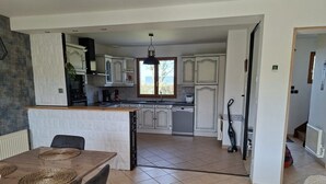 Een ruime keuken met witte kastjes, een eethoek met een houten tafel en stoelen, en een doorkijk naar een zithoek.