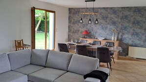 Een moderne woonkamer met een grijze bank, een eethoek met een tafel en stoelen, en een groot raam met uitzicht op groen.