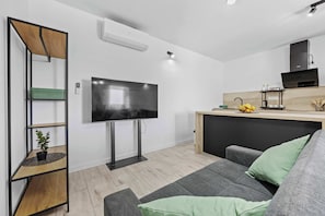 A modern living room with a flat-screen TV, a grey sofa, a wooden shelf unit, and a kitchen area with a black countertop and wooden cabinets.