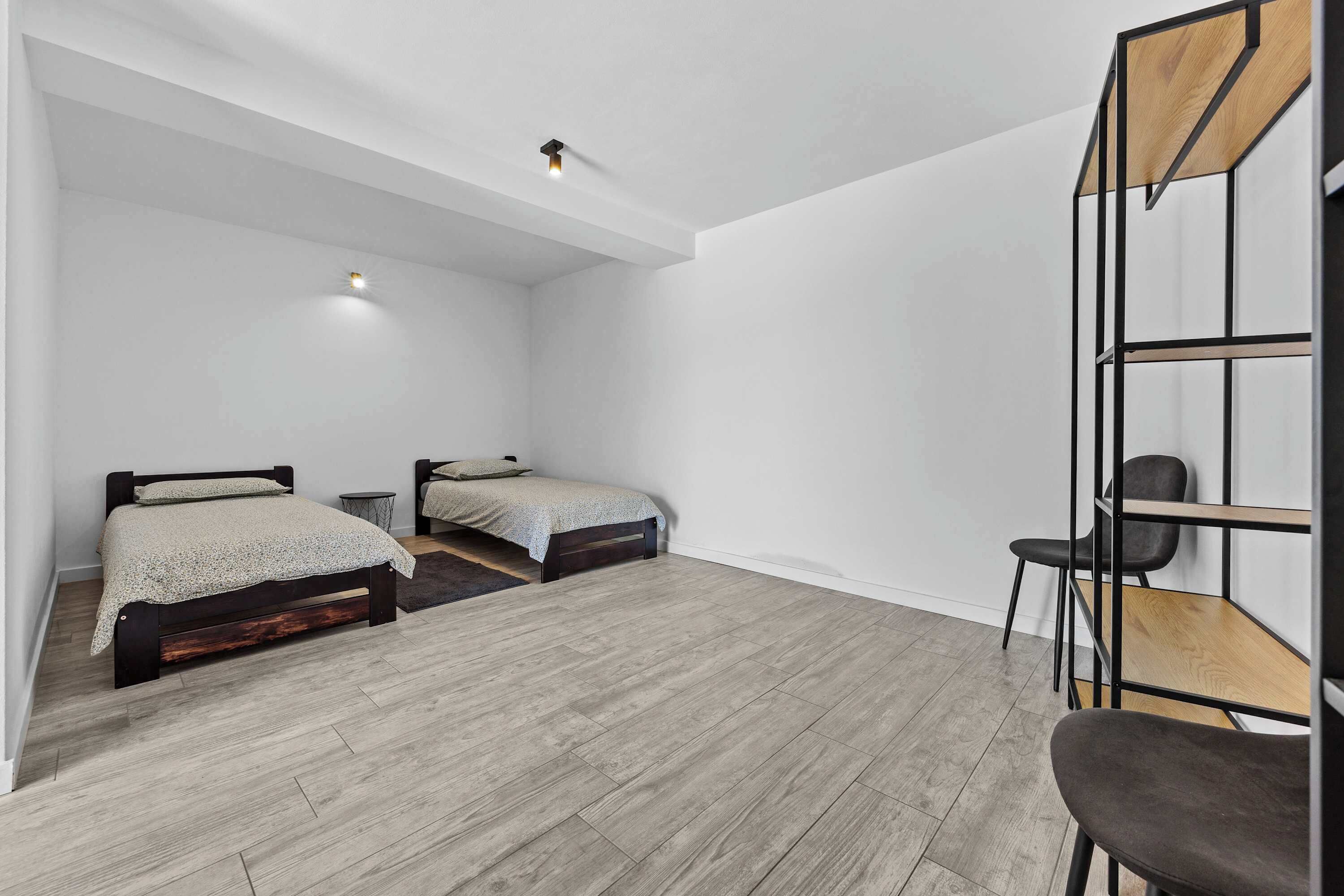 A room with two beds, a chair, a desk, and a shelving unit.