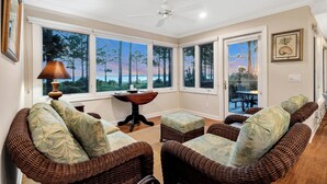 A room with wicker furniture, large windows, a ceiling fan, and a table with a lamp.