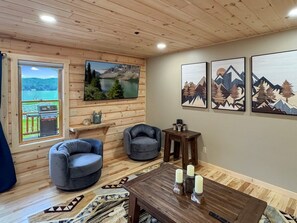 A cozy living room with a wooden ceiling, a large window with a view, two grey armchairs, a coffee table with candles, and framed artwork on the wall. - Townhome O3 - Luxury Waterfront Home on 4th Lake (Inlet)