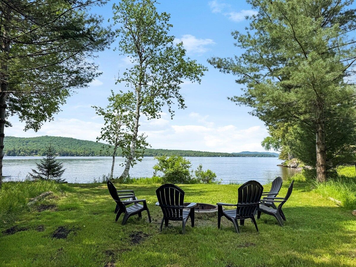 An outdoor seating area with chairs and a table, overlooking a lake with trees and hills in the background.
