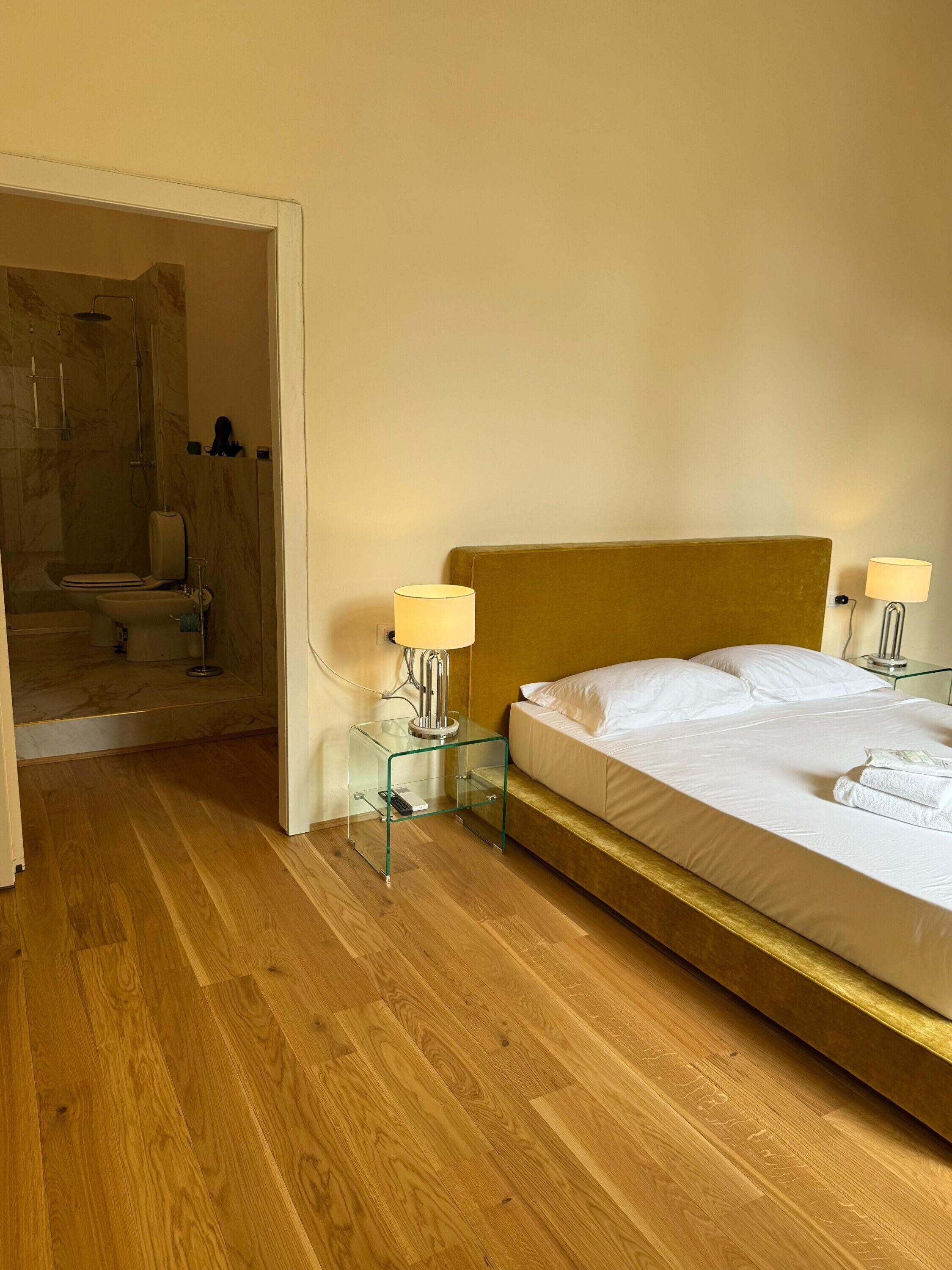 A hotel room with a wooden floor, a bed with white linens, two bedside lamps, and a glass side table.
