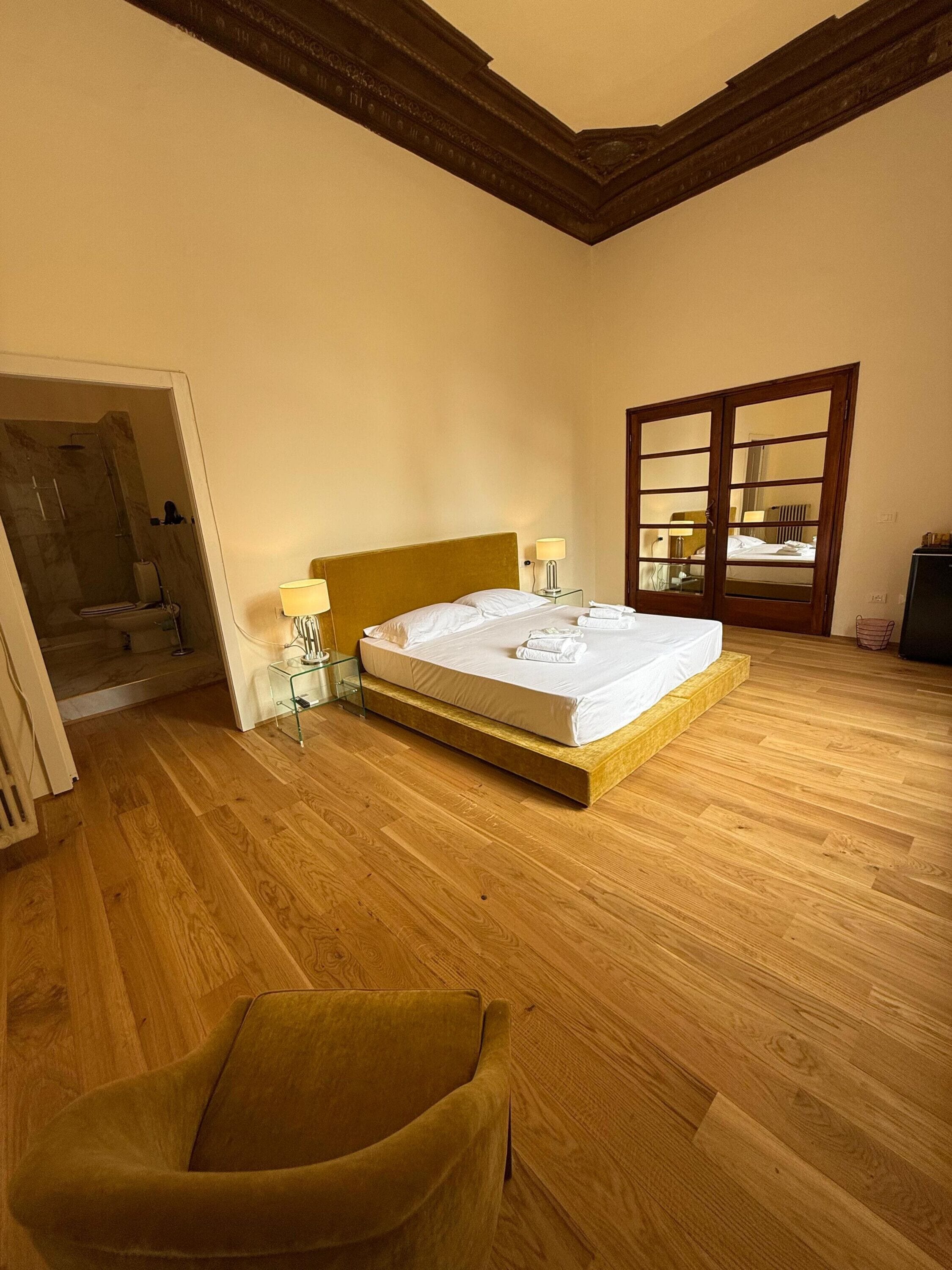 Un dormitorio con suelo de madera, una cama con ropa blanca, cabecero de madera, mesita de noche de cristal y un sillón amarillo, además de una puerta con paneles de vidrio.