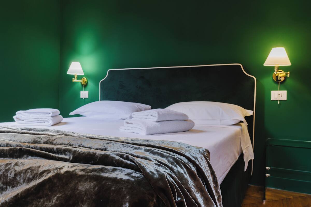 A neatly made bed with white linens and a dark headboard in a room with green walls and two wall-mounted lamps.