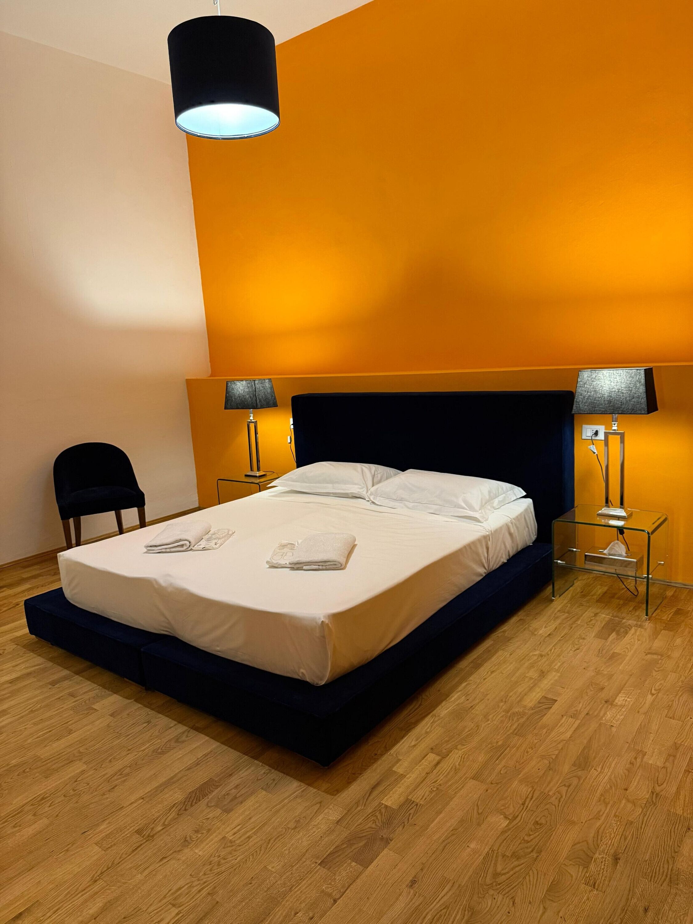 A neatly made bed with white linens and two pillows, flanked by two bedside tables with lamps, in a room with wooden flooring and a dark blue headboard.