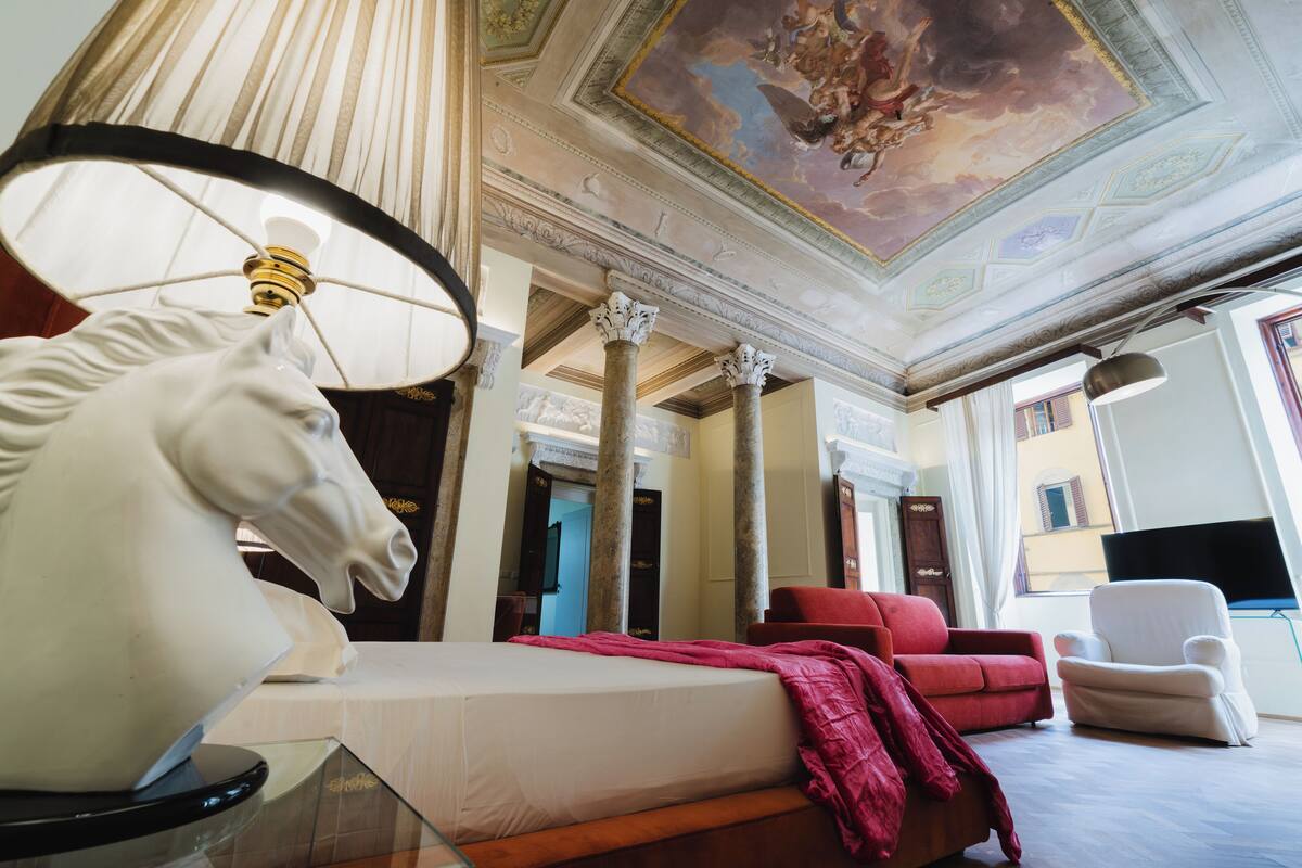 A hotel room with a bed, a red sofa, a white armchair, a ceiling with a fresco, and a large window.