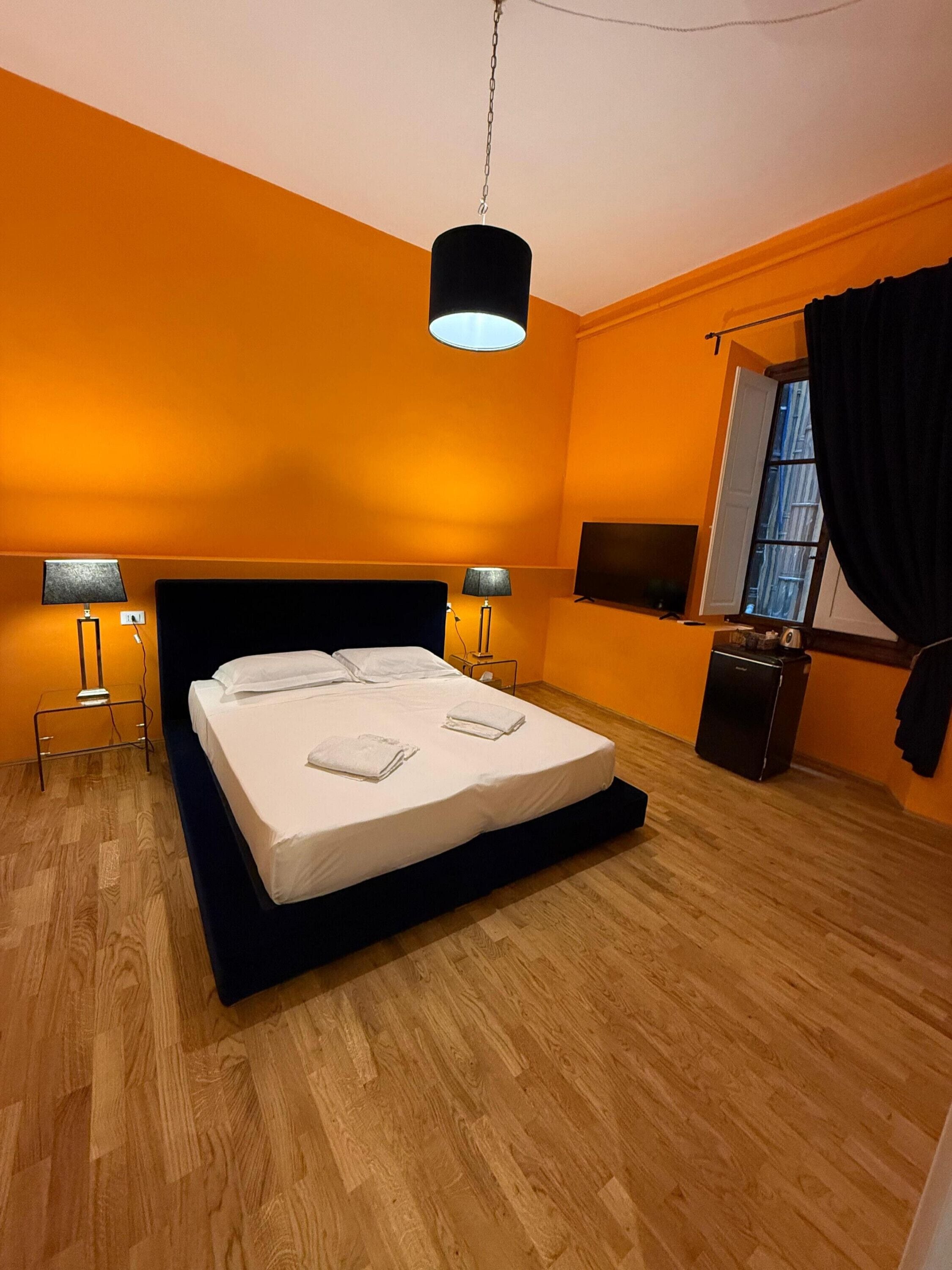 A modern hotel room with a large bed, two bedside tables with lamps, a flat-screen TV, and a small refrigerator.