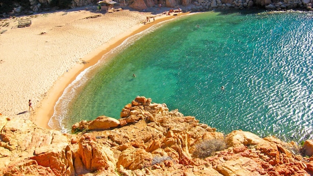 A coastal beach with clear turquoise waters, rocky cliffs, and a sandy shore.