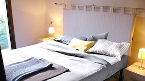 A neatly made bed with a grey and white duvet, a blue throw blanket, and several pillows. There is a bedside table with a lamp and a small drawer. The room has a window with a view of greenery outside.