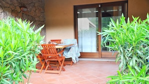 A patio with a wooden table and chairs, a stone wall, and a glass door leading to a covered area.