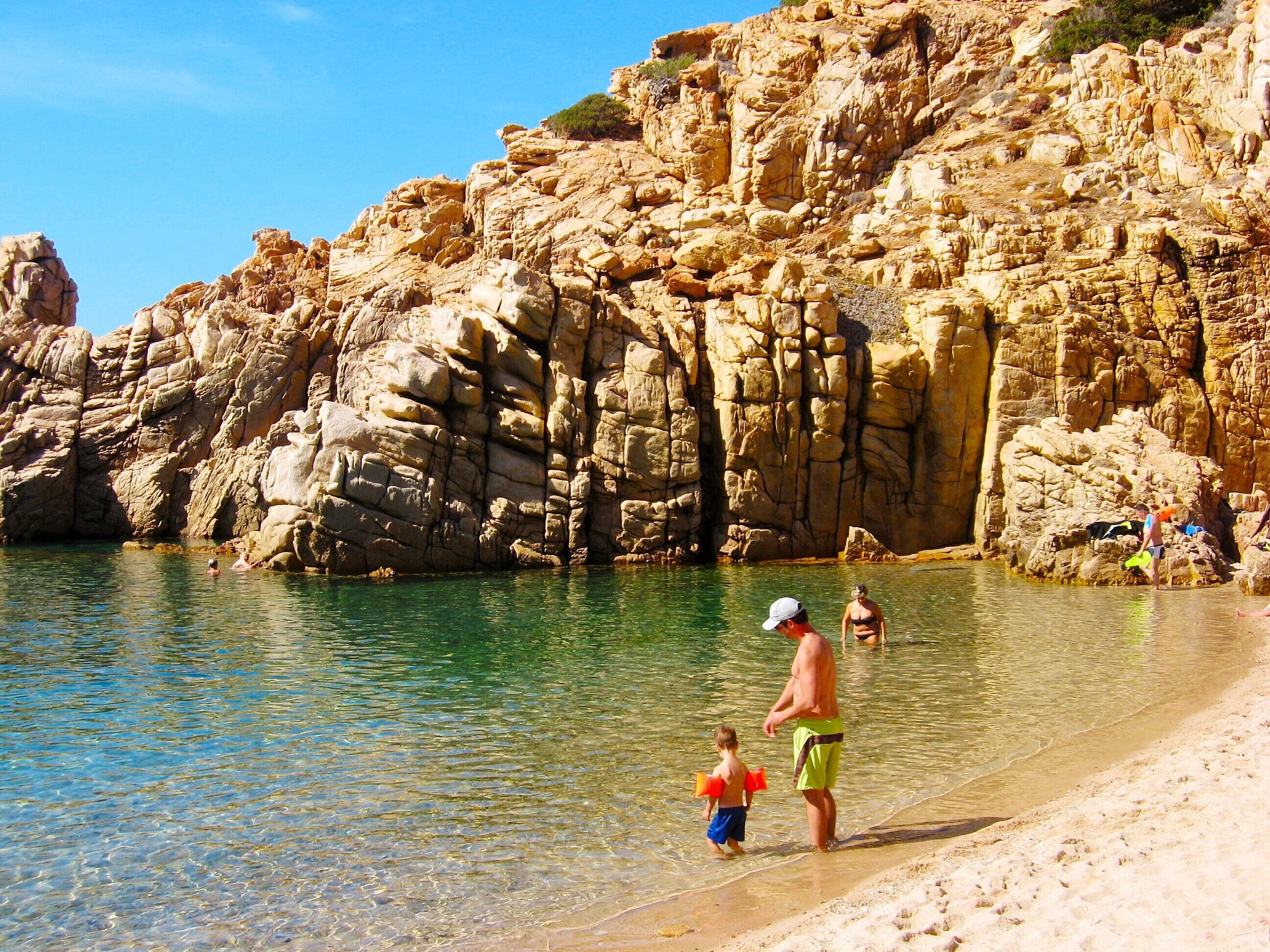 A rocky coastline with clear water, a sandy beach, and people swimming and walking.