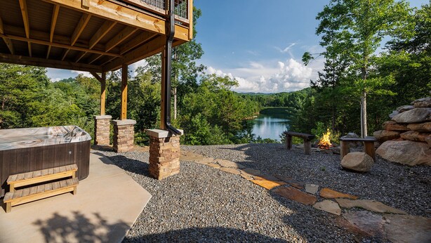 Una terraza de madera con jacuzzi, pilares de piedra y una chimenea, con vistas a un lago.