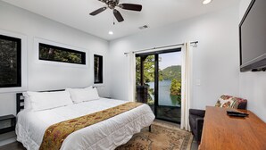 Un dormitorio con cama, ventilador de techo, ventana con vista a un lago y una mesa de madera con control remoto.
