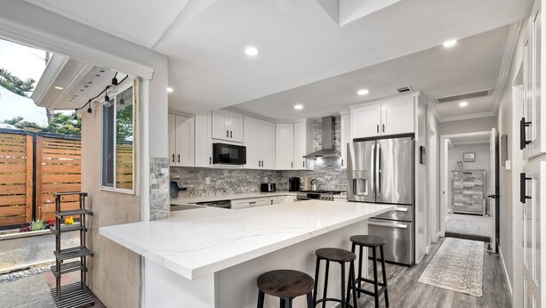 Une cuisine moderne avec des meubles blancs, un grand îlot central, des appareils électroménagers en acier inoxydable et une vue sur l’extérieur.