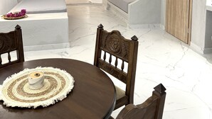 Une pièce moderne et minimaliste, avec un plafond blanc et incurvé et une porte en bois. Une table ronde, surmontée d’un set de table en fibres tressées et d’un petit vase, se trouve au premier plan.