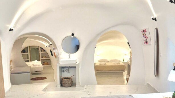 Une chambre avec un lit, un miroir rond, un lavabo et une petite table. Deux arcades mènent à une autre pièce.