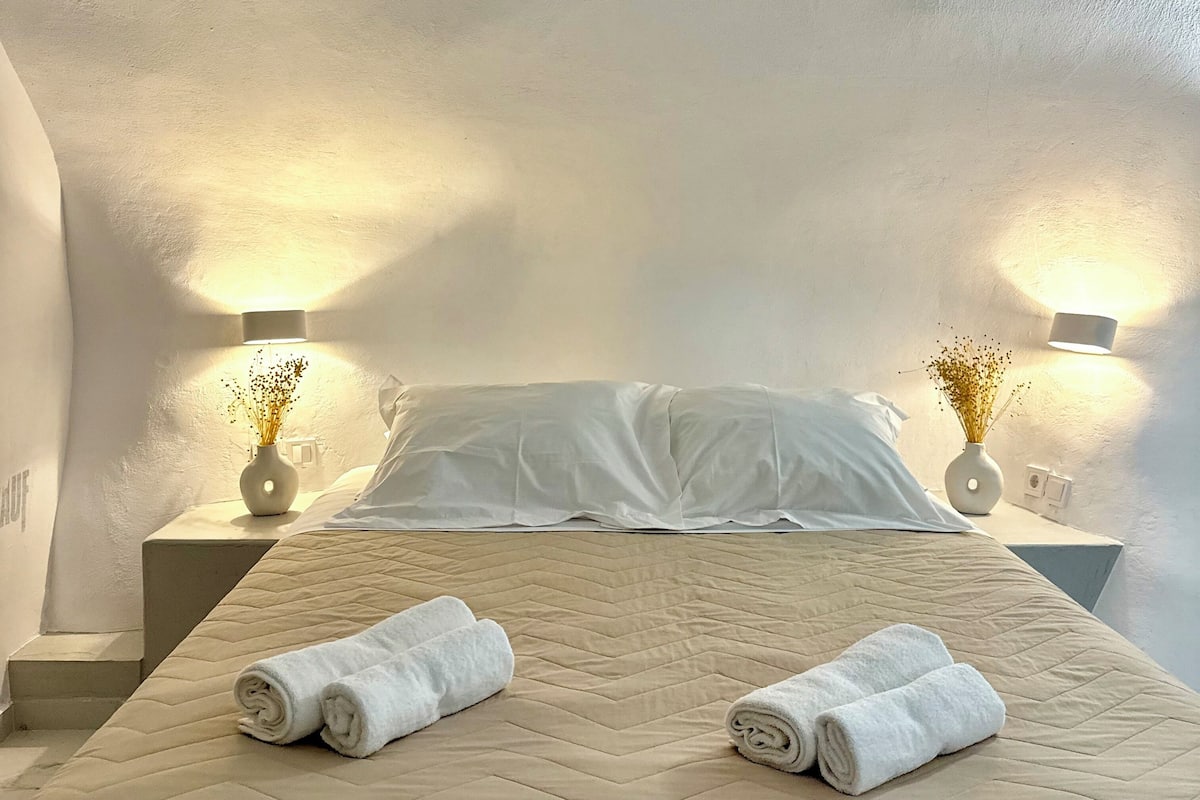 A neatly made bed with two rolled towels, a beige quilted bedspread, and two wall-mounted lamps with vases on bedside tables.