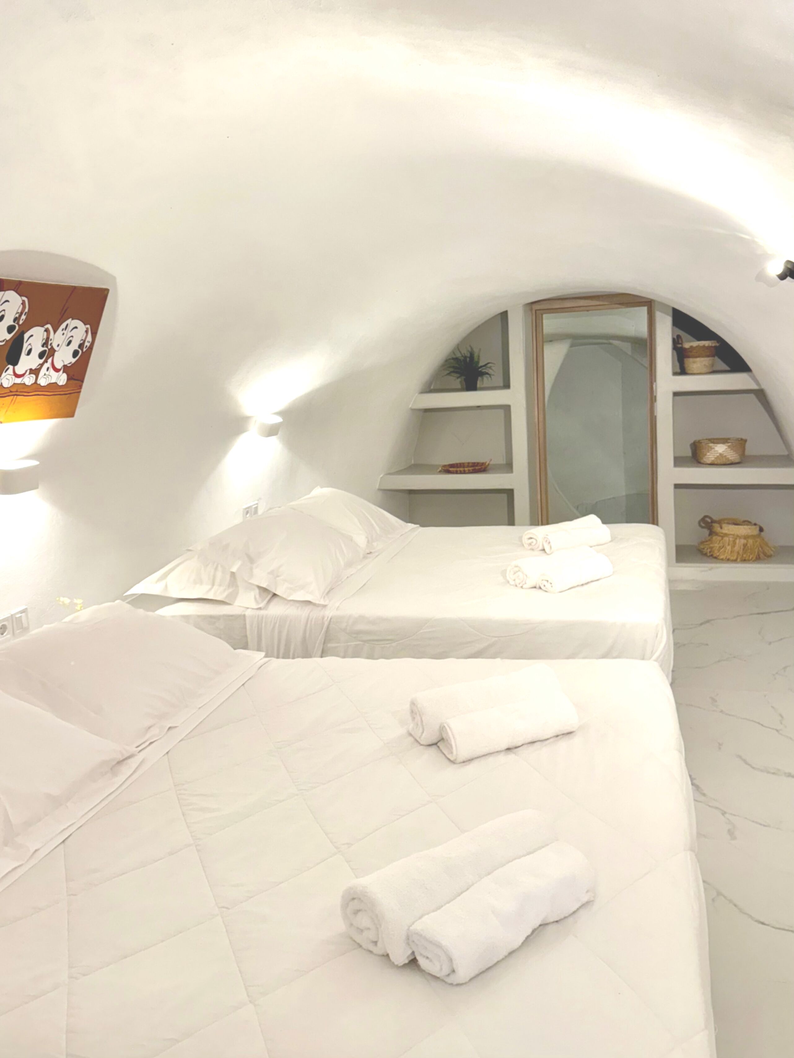 A white, arched room with two beds, a mirror, and shelves with decorative items.