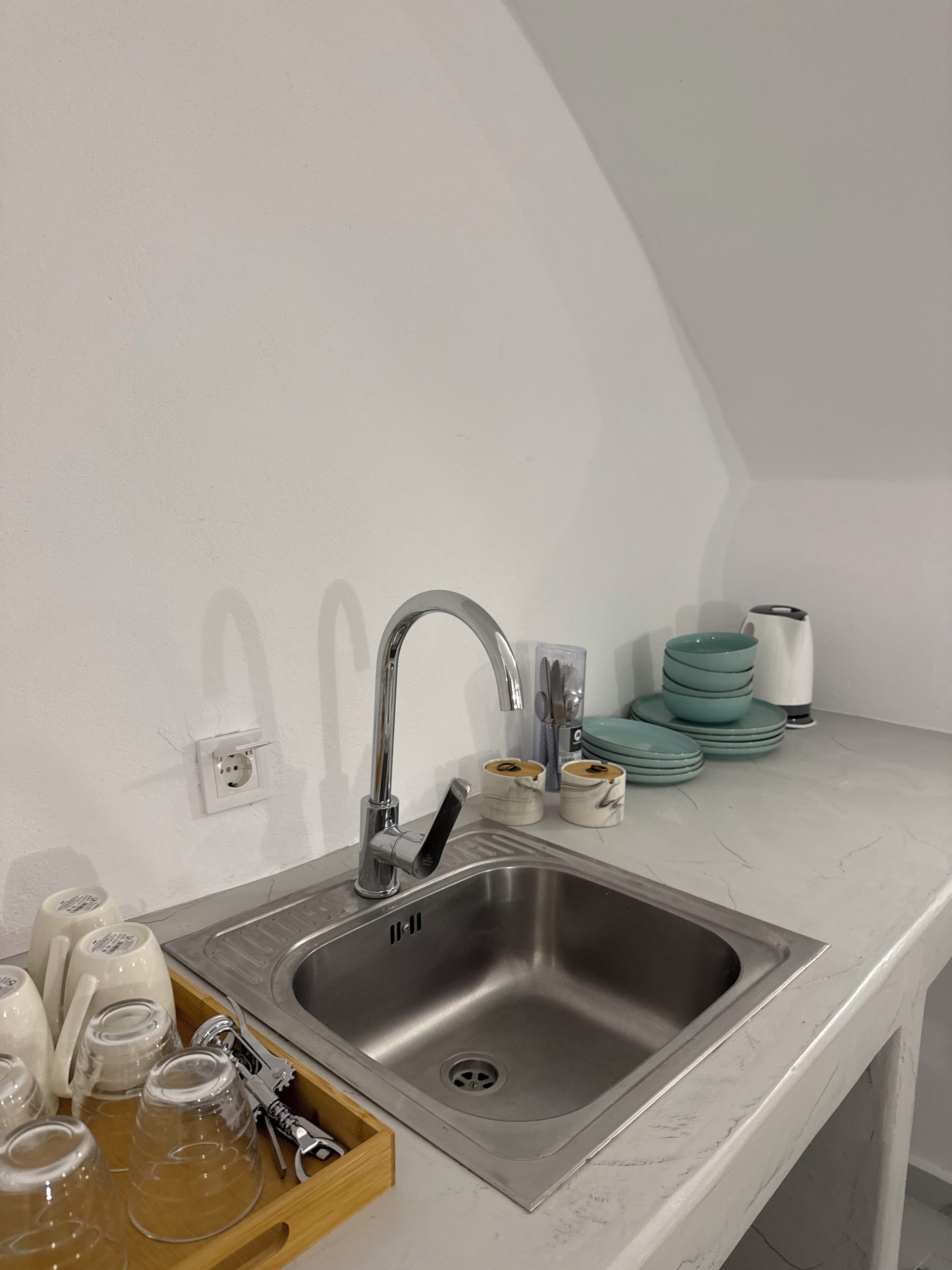 A modern kitchen with a stainless steel sink, a white countertop, and a wooden tray with glasses and utensils.