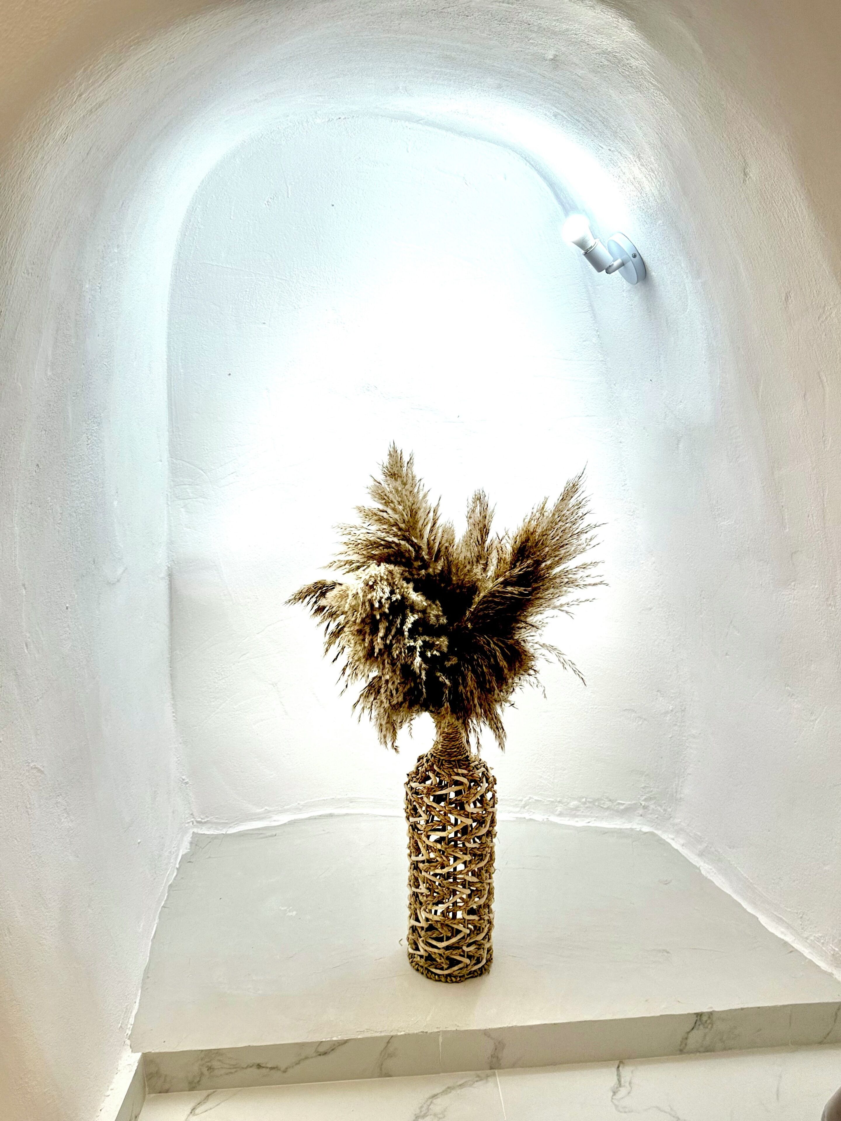 Rincón con pared blanca y base de mármol, que presenta un jarrón decorativo tejido con plantas secas.