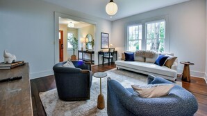 Una sala moderna con sofá, sillones y una mesa de centro. Se observa un comedor con mesa y sillas, y una cocina al fondo.