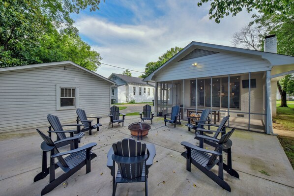 Un patio con sillas Adirondack dispuestas alrededor de un brasero, una zona de estar cubierta y una vista de una casa al fondo.